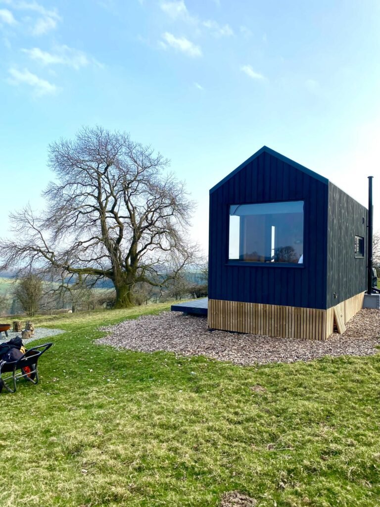 Black cabin in the countryside, with trees, wheelbarrow and fireplace