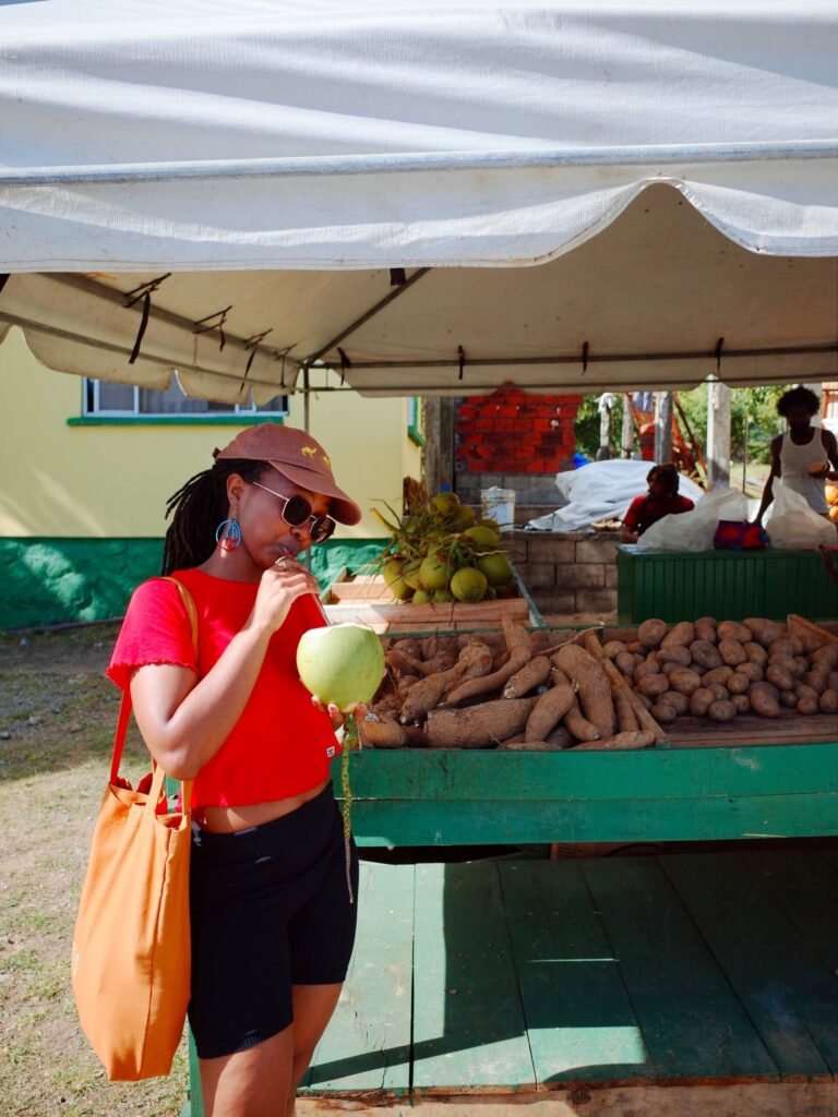 Girl wearing red top and black shorts, sipping fresh coconut water. Market stall in the background with yams, potatoes and caribbean people