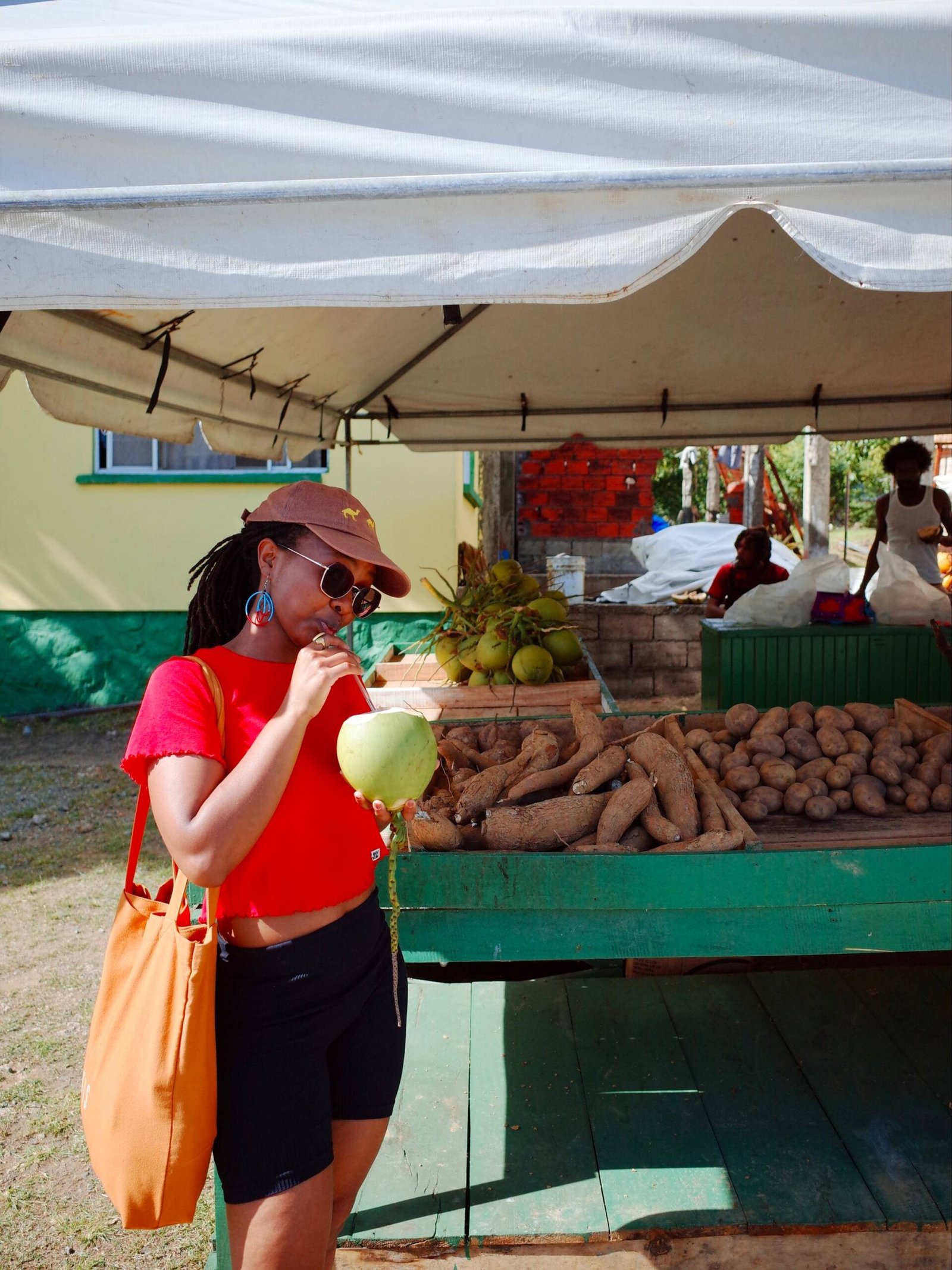 Girl wearing red top and black shorts, sipping fresh coconut water. Market stall in the background with yams, potatoes and caribbean people