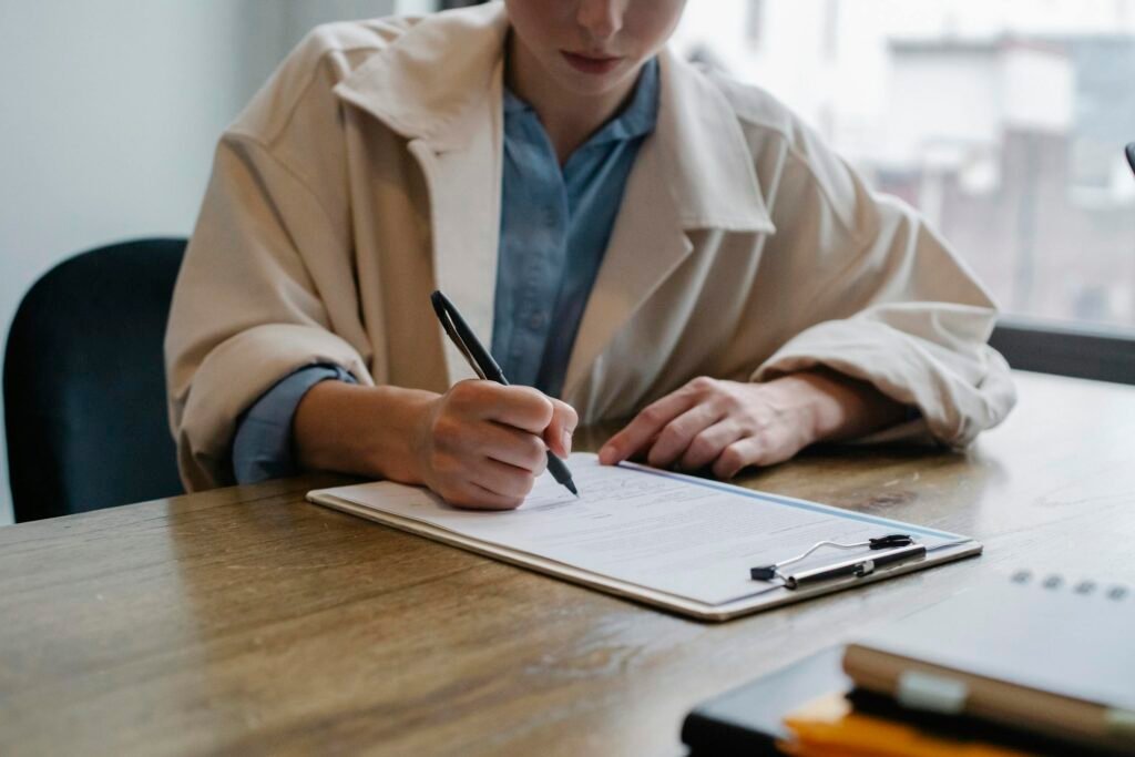 A woman in a formal setting fills out visa paperwork on a clipboard at an office desk.