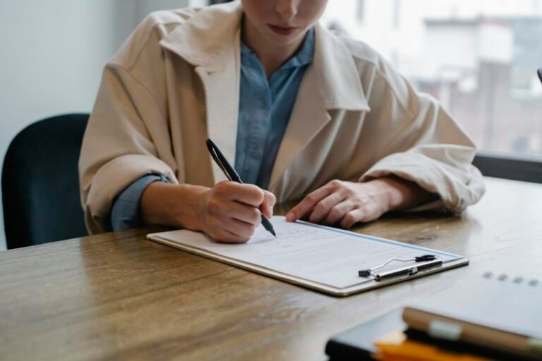 A woman in a formal setting fills out visa paperwork on a clipboard at an office desk.