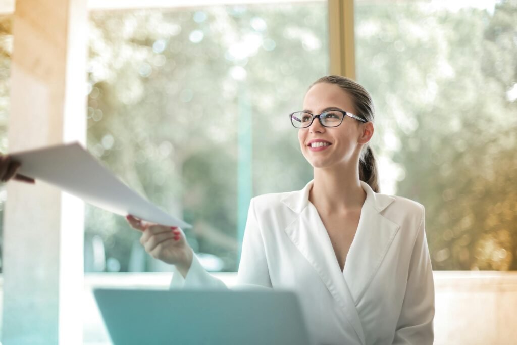Cheerful woman sitting near a laptop passing documents, presumably visa sponsorship documents, to a colleague